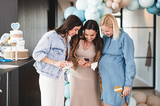 A happy group of female close friends looking at a cute polaroid photo they have taken together at a baby shower. The girls are smiling while observing the photo. They are dressed in color-coordinated clothes to celebrate their best friend is having a boy. Beautiful baby shower celebration with cute blue decorations at a modern apartment.