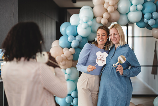 A young Caucasian mom-to-be and her female best friend taking photos together with funny photo props at a baby shower. Their black friend is taking a photo with a polaroid camera. They are glowing and look happy to be making such fun memories together.