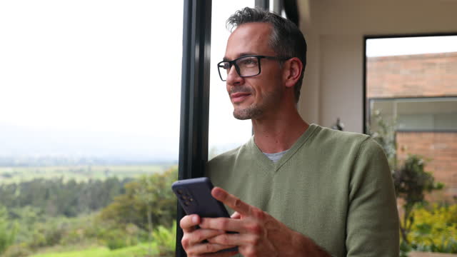 Handsome Latin American man at home leaning against the window texting on smartphone and looking away slightly smiling