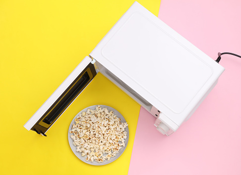 Microwave oven with a bowl of popcorn on yellow-pink pastel background. Top view