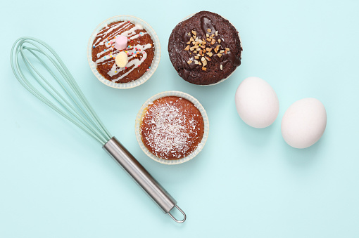 Cooking concept. Cupcakes with raw eggs and whisk on a blue background. Top view