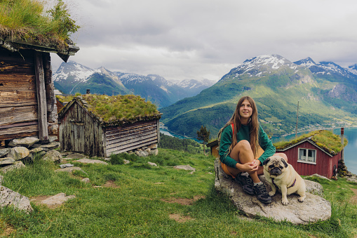Portrait of smiling female backpacker and a cute dog sitting on the stone on the top of Rakssetra and enjoying a view of the old viking village, mountains and the fjord in Stryn, Vestland country