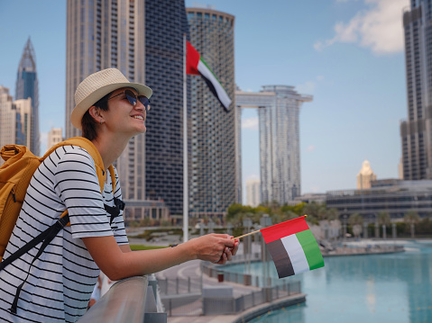 Enjoying travel in United Arabian Emirates. Young woman with yellow backpack with the flag of the United Arab Emirates walking on Dubai Downtown in sunny summer day.