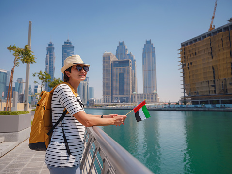 asian happy traveler woman with hat and yellow backpack with the flag of the United Arab Emirates enjoys stunning panoramic view of Dubai Creek Canal and famous tallest skyscraper Burj Khalifa