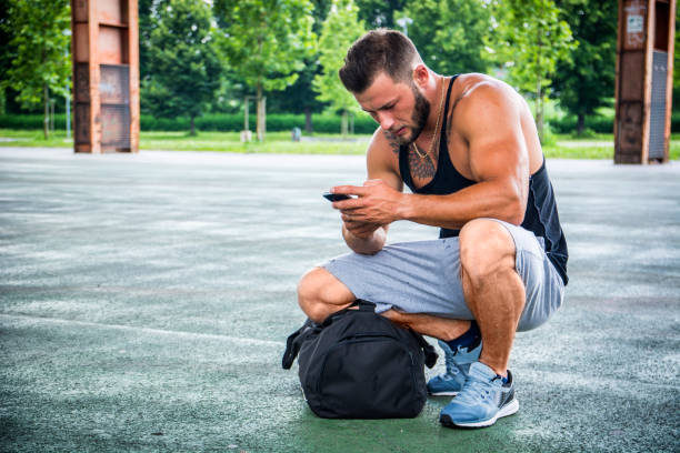 homme musclé attrayant utilisant un téléphone portable dans un parc de la ville - crouching male men sensuality photos et images de collection