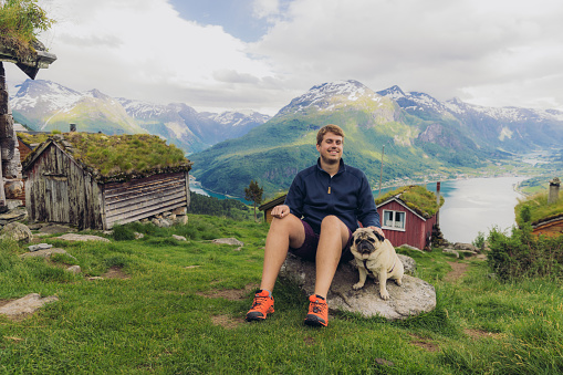Smiling male relaxing with a dog in the mountains with view of authentic viking town and the fjord from above in Rakssetra