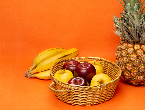 Still life of healthy fruits of yellow color apples, red color apples in a basket , bananas and exotic tropical pineapple fruit on orange color background