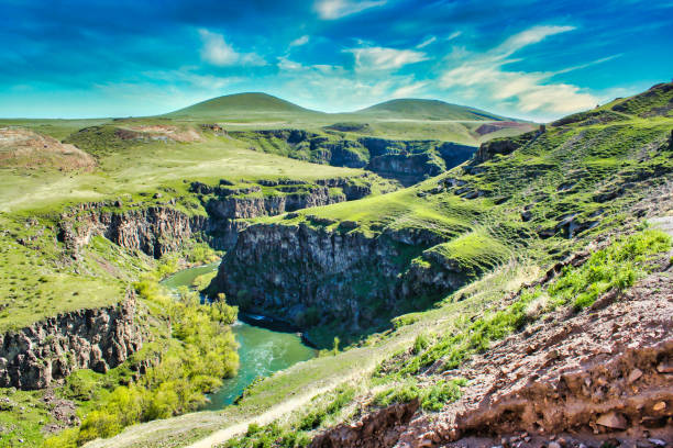 A wide shot of a river valley near the ancient ruins of Ani city in Kars, Turkey. The landscape is gorgeous with the curves of the river and the slopes of the surrounding green hills. The altitude is approximately 2400 meters and there are almost no trees visible on the hills. There are only  trees by the side of the water.  The valley is curved and gets deeper into the scenery.  There are two hills in the background. The sky is blue with some clouds. The shot is taken in springtime in the province of Kars, the very east of Turkey near the Armenian border.