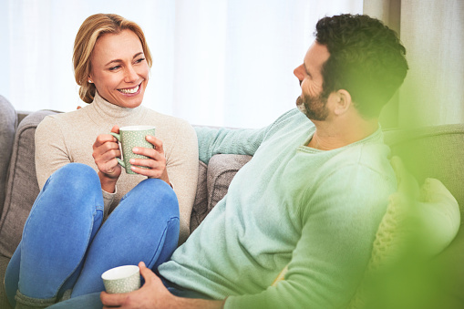 Couple, coffee and happy in living room on sofa, bonding and conversation in home. Tea, smile and mature man and woman relaxing, talking and enjoying quality time together on couch in lounge of house