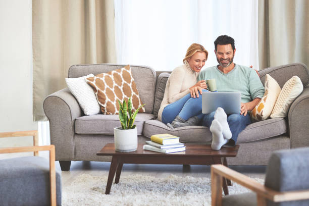 Coffee, couple and happy with laptop in living room of home, bonding and relaxing. Tea, couch and man and woman with computer, watching and streaming video, film or movie together on sofa in house. stock photo