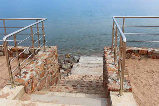 Marble stone staircase straight into the blue sea with handrails and sand. Coastline. Summer. Copy space