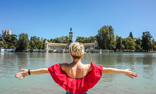 Happy woman with outstretched arms in front of the lake and Alfonso XII Monument in the Buen Retiro Park (Parque del Buen Retiro) in Madrid, Spain