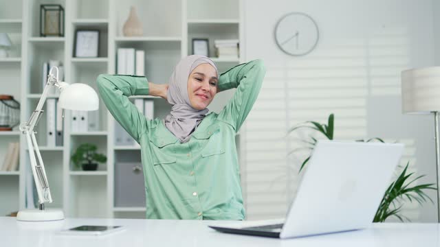 A satisfied glad young female muslim worker employee relaxes after finishing work on a laptop computer
