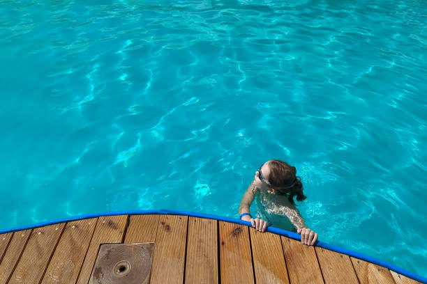 Girl is wearing swimming goggles in the pool. She looks to the right and holds her hands on the pool side outdoors