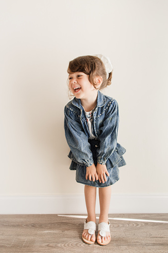 A Cute & Sassy 4-Year-Old Cuban-American Toddler Girl with Brown Eyes & Brown Curly Hair in a Large Retro-Style Scrunchie While Wearing Her Mother’s Vintage 1990’s Denim Jacket & Skirt Outfit with White Sandals in a Neutral Indoor Space with Copy Space