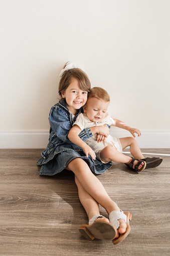 A 4-Year-Old Cuban-American Girl with Brown Eyes & Curly Hair Wearing a Vintage 1990’s Denim Outfit While Sitting with Her 16-Month-Old Brother Wearing a Cream Linen Romper & Brown Leather Sandals Indoor in a Neutral Clean Room with Natural Light