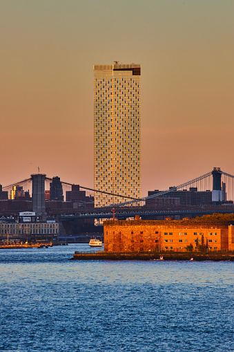 Image of Lone skyscraper and Governors Island with Manhattan and Brooklyn Bridges from water in golden hour of New York City