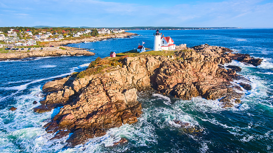 Image of Waves crash over rocks aerial over Maine island with lighthouse and view of homes on mainland