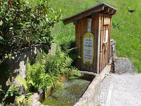 Ottenhoefen, Germany – July 08, 2022: An outdoor outhouse is situated near a bubbling fountain, surrounded by lush green foliage