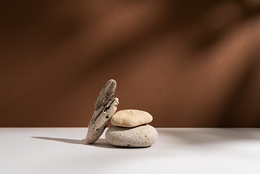 Empty stone podium with lights and shadows on brown background. Minimal backdrop. Round natural rock and piece of driftwood. Abstract pedestal or showcase for presentation. Minimal wabi sabi concept.