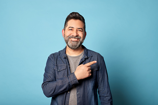 Mid adult latin man smiling pointing to the side showing copy blank space, isolated over blue background. Indoor studio shot. Advertisement & presentation concept. Copy space.
