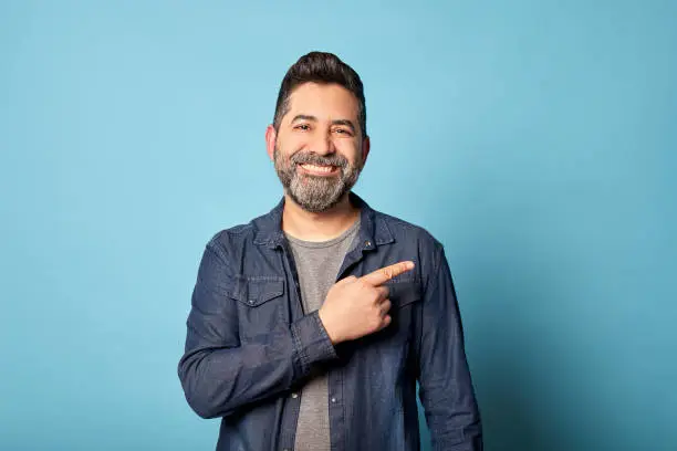 Photo of Mid adult latin man smiling pointing to the side showing copy blank space, isolated over blue background. Indoor studio shot. Advertisement & presentation concept. Copy space.