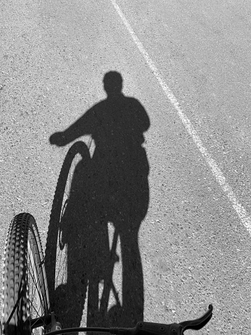 The image has part of the bicycle visible in the lower left corner, the shadow of the silhouette of the cyclist and his bike emerges from the bike tire, limited by the line separating the tracks on the road\nblack and white photo