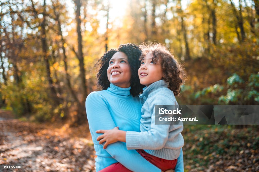 Good Looking Mixed Race Mother And Son Amazed By The Beauty Of Nature Cheerful multicultural mother and son enjoying nature. They are outdoors in the forest. The mother is holding her son in her arms. Autumn Stock Photo