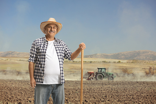 Mature farmer with a shovel posing on a field with a tractor operating in the back