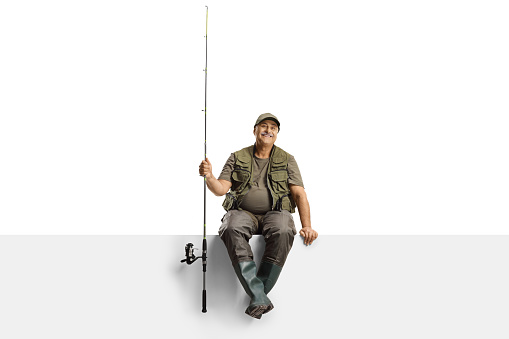 Mature fisherman with a fishing rod sitting on a blank panel isolated on white background