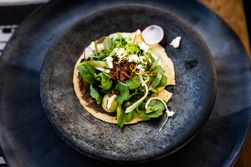 A horizontal overhead photograph showcasing a traditional pre-Hispanic dish from Oaxaca, Mexico. The dish features a mole tostada topped with chapulines (grasshoppers) and quelites (greens) served on a black clay plate from Oaxaca. The presentation is set against a rustic wooden table background, emphasizing the authenticity and cultural roots of this unique Mexican culinary experience.