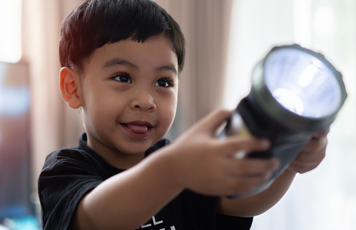 Asian toddler in black costume shines a flashlight forward in the room and smiles with joy at what he sees. Close-up