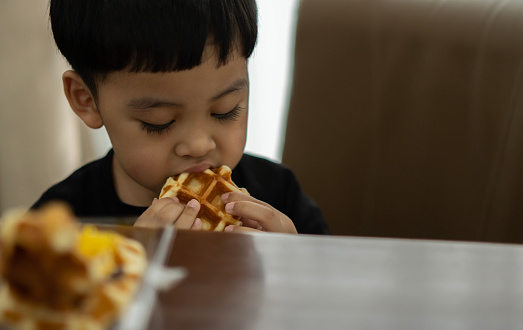 Portrait of happy Asian little preschool boy holding fresh baked heart waffle. Kids eating a waffles. Top view.