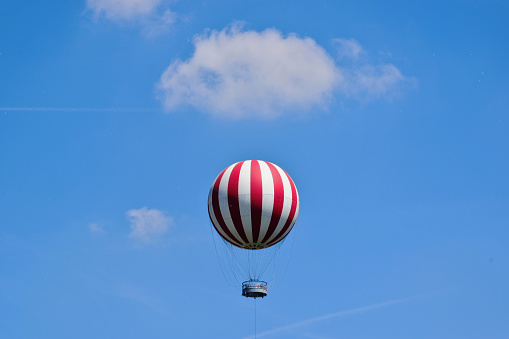 isolated gas filled red and white observation balloon. tourist attraction. round passenger basket. blue sky background with white clouds. leisure and outdoors