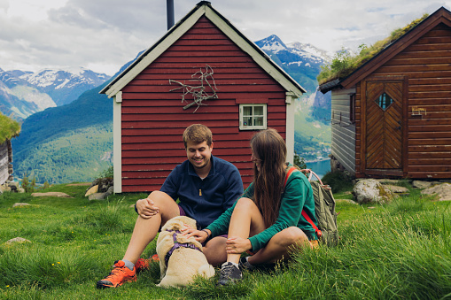 Smiling female and male sitting with a dog in the mountains with view of authentic viking town and the fjord from above in Rakssetra, Stryn, Western Norway