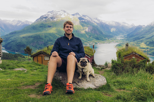 Portrait of smiling male sitting with a dog in the mountains with view of authentic viking town and the fjord from above in Rakssetra, Stryn