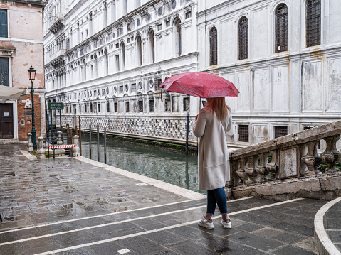 Venice, Italy - April 24, 2023: High resolution. Young woman taking photos in the rain with her smartphone of the Doge's Palace and the Bridge of Sighs
