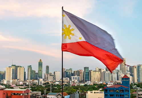 In the early evening afterglow of sunset,the red,blue and white Philippine flag flies,with it's golden sun and stars showing, in the warm breeze, on a city rooftop,tall modern buildings behind.