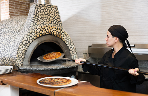 Focused young adult woman chef working in pizzeria, holding freshly oven baked pizza on paddle