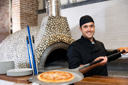 Portrait of smiling pizza chef at work, taking pizza from oven at restaurant kitchen