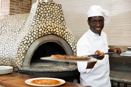Positive young adult man working in pizzeria, holding big shovel with freshly baked pizza