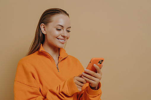 Happy charming young woman holding smartphone in hands and smiling, chatting in social networks, pleasant female in orange sweatshirt using mobile phone while standing isolated on beige background