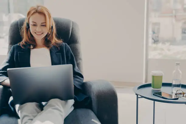 Happy smiling female freelancer during online conference via modern laptop, cheerful redhead businesswoman in headset watching webinar or educational video while relaxing on armchair at home
