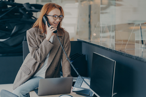 Woman in spectacles working in office wearing casual clothes, talking on phone while sitting on her desk, modern and comfortable office environment, blurred background. Happy employee at work concept