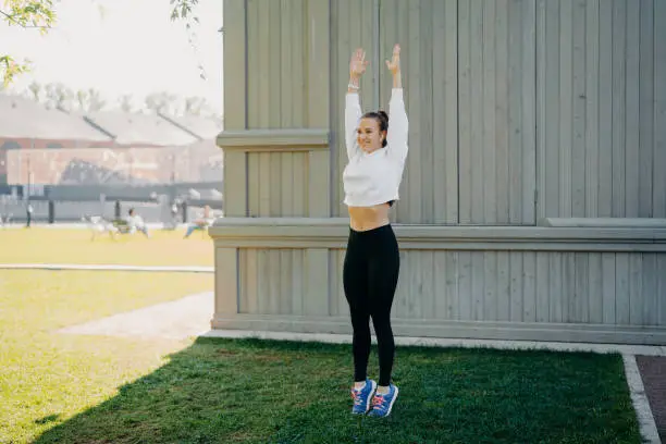 Athletic slim brunette woman jumps and does sport exersices outdoors keeps arms raised up has work out in morning wears white sweatshirt leggings and sportshoes poses in full length on green lawn