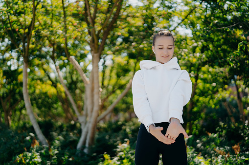 Horizontal shot of sporty European woman stretches hands and warms up before workout wears white sweatshirt andleggings poses against trees background spends free time outdoors on fresh air.