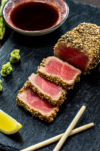 Vertical close up of appetizing sesame crusted tuna steak. Medium rare Yellow fin tuna slices on black slate with wasabi, soy sauce and chopsticks.