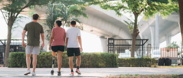 Woman exercising in a park with a friend providing support while using a prosthetic leg. People jogging side by side outside in a park. Female walking and exercise works out outside.