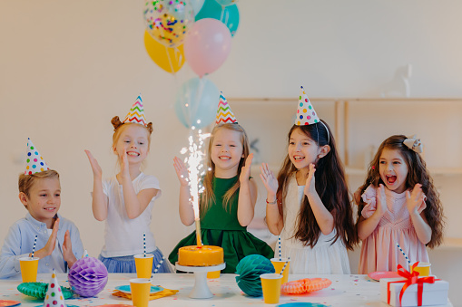 Overjoyed children friends clap hands and looks at big sparkle on cake, stand near festive table with present, cups and holiday attributes, have fun together, celebrate birthday