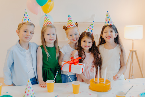 Glad birthday girl stands with presenet, happy friends come to congratulate her, wear party cone hats, stand near festive table with cake, smile joyfully, celebrate festive event